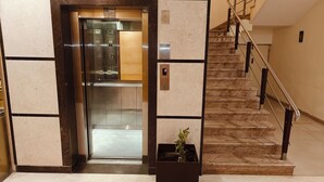 Een lift met een digitaal scherm, een bedieningspaneel en een bordje dat aangeeft dat hij buiten gebruik is. Naast de lift bevindt zich een trap met een leuning en een bloempot.