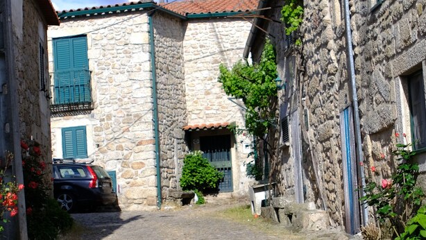 A cobblestone street flanked by stone buildings with green shutters and a parked car. - 9, Cimo da aldeia (Aldeia de João Pires)