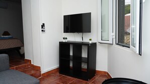 A modern room with a flat-screen TV mounted on a black console table, a bed with a quilted cover, a grey sofa, and a window with a view of a stone wall and greenery. - 9, Cimo da aldeia (Aldeia de João Pires)