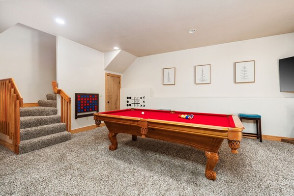 Een pooltafel in een kamer met vloerbedekking, een houten trap en beelden in lijsten aan de muur.
