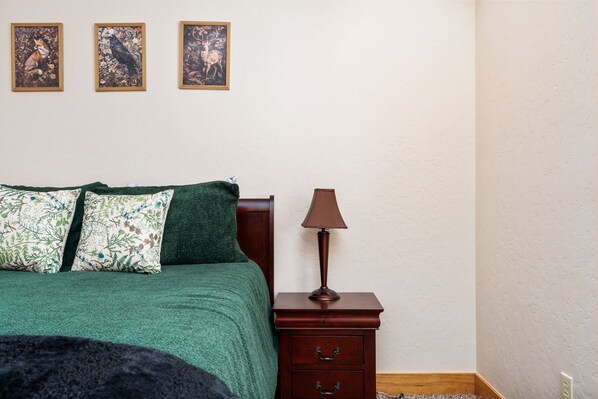 Un dormitorio con una colcha verde, dos almohadas, una mesita de noche de madera con una lámpara y tres cuadros en la pared.