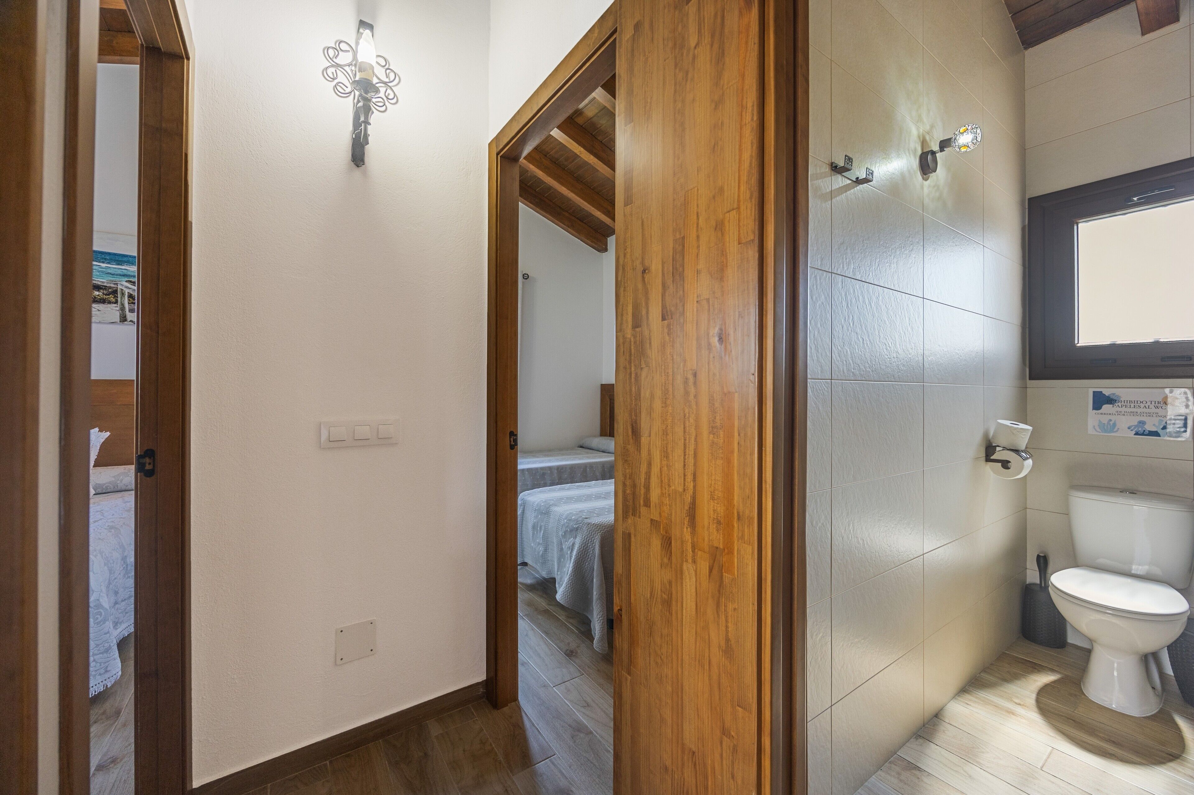 A hallway with a wooden door, a bed with a white bedspread, a white toilet, and a window.