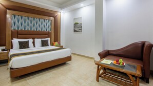 Une chambre d’hôtel avec un grand lit, un canapé en cuir marron, une petite table sur laquelle se trouve un panier de fruits, et un tableau accroché au mur.