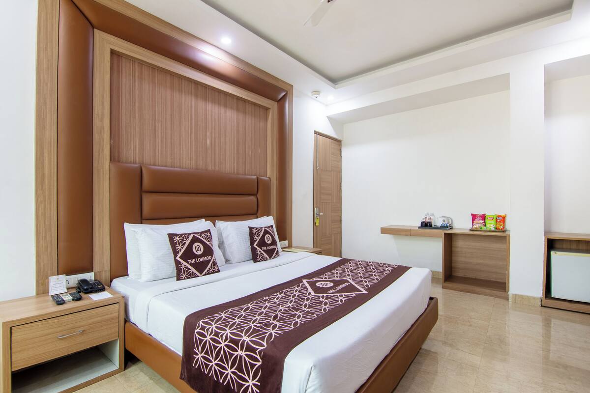 A hotel room with a large bed, wooden headboard, a bedside table with a phone, and a small shelf with toiletries.