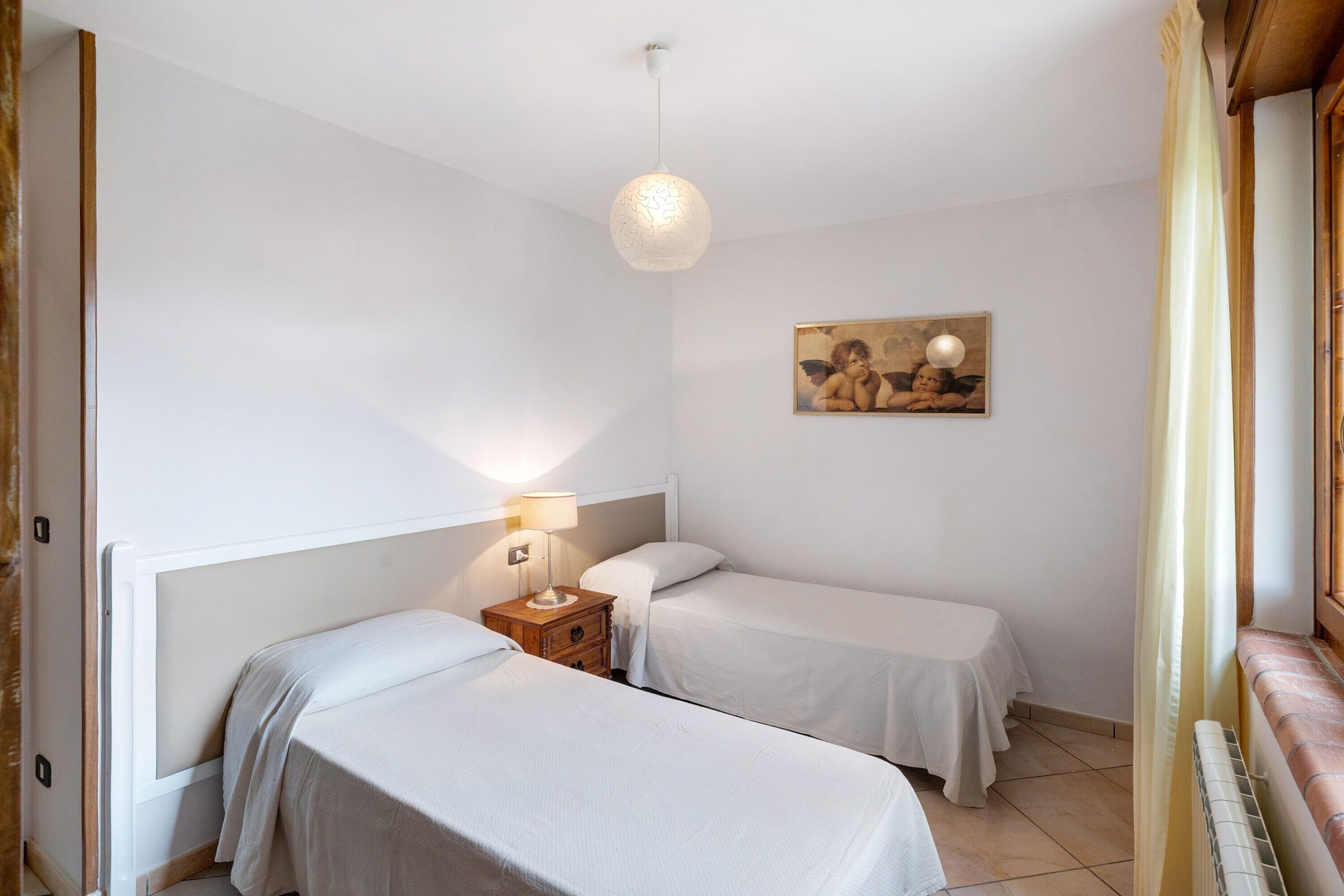 A room with two beds, a nightstand with a lamp, a painting on the wall, and a window with curtains.