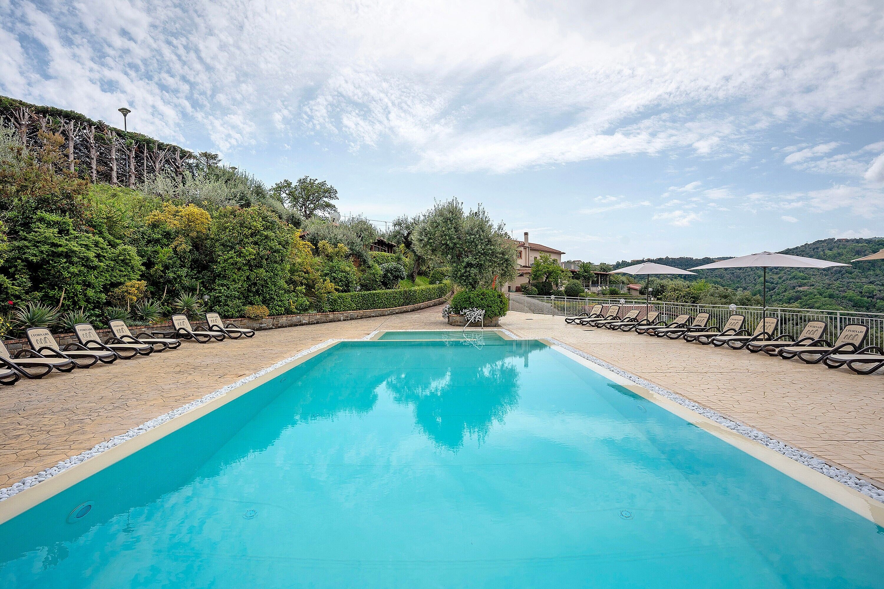A swimming pool with lounge chairs and umbrellas, surrounded by greenery and hills.