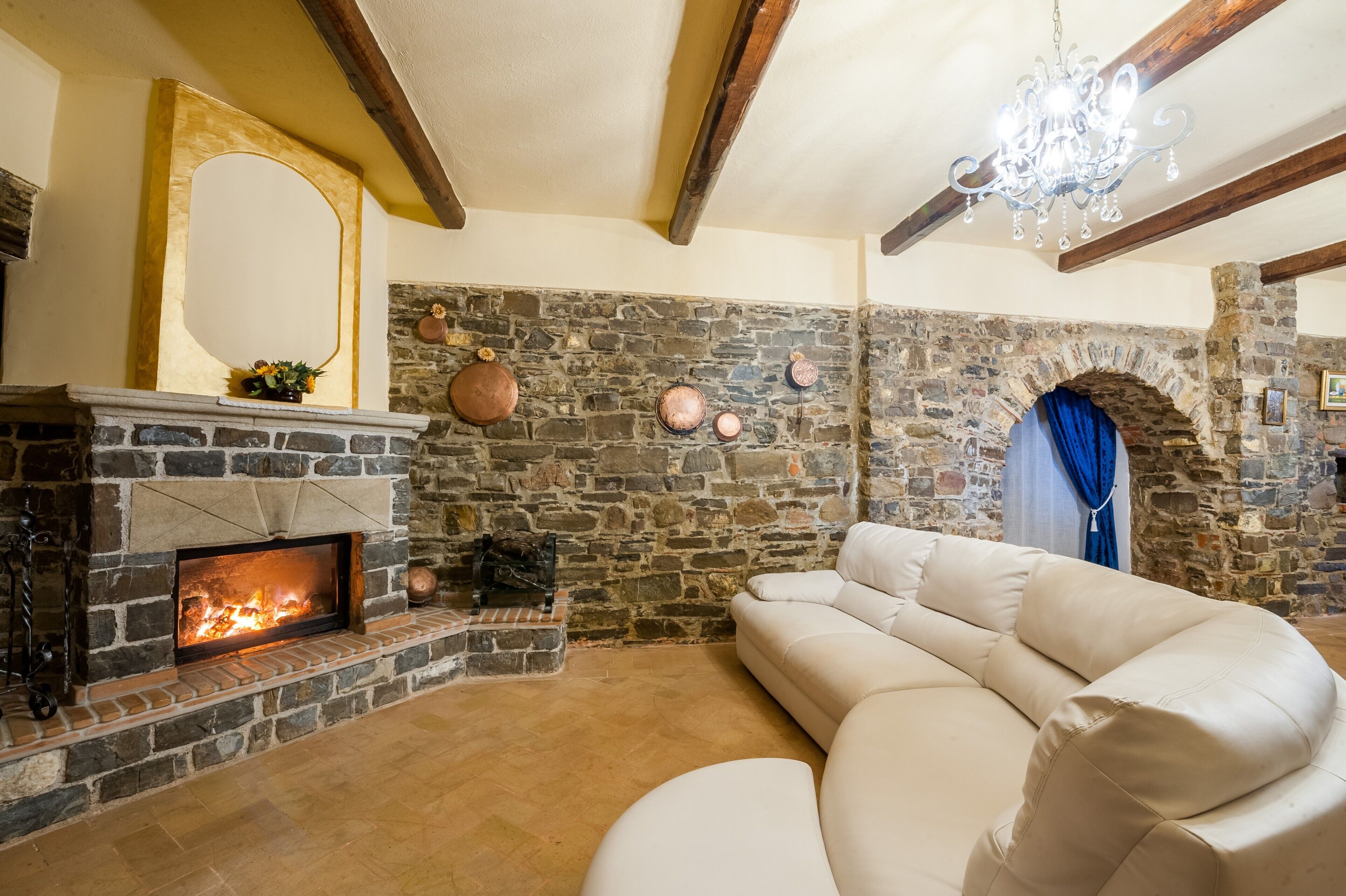 Une cheminée en pierre avec un feu de bois, un canapé beige et un lustre.