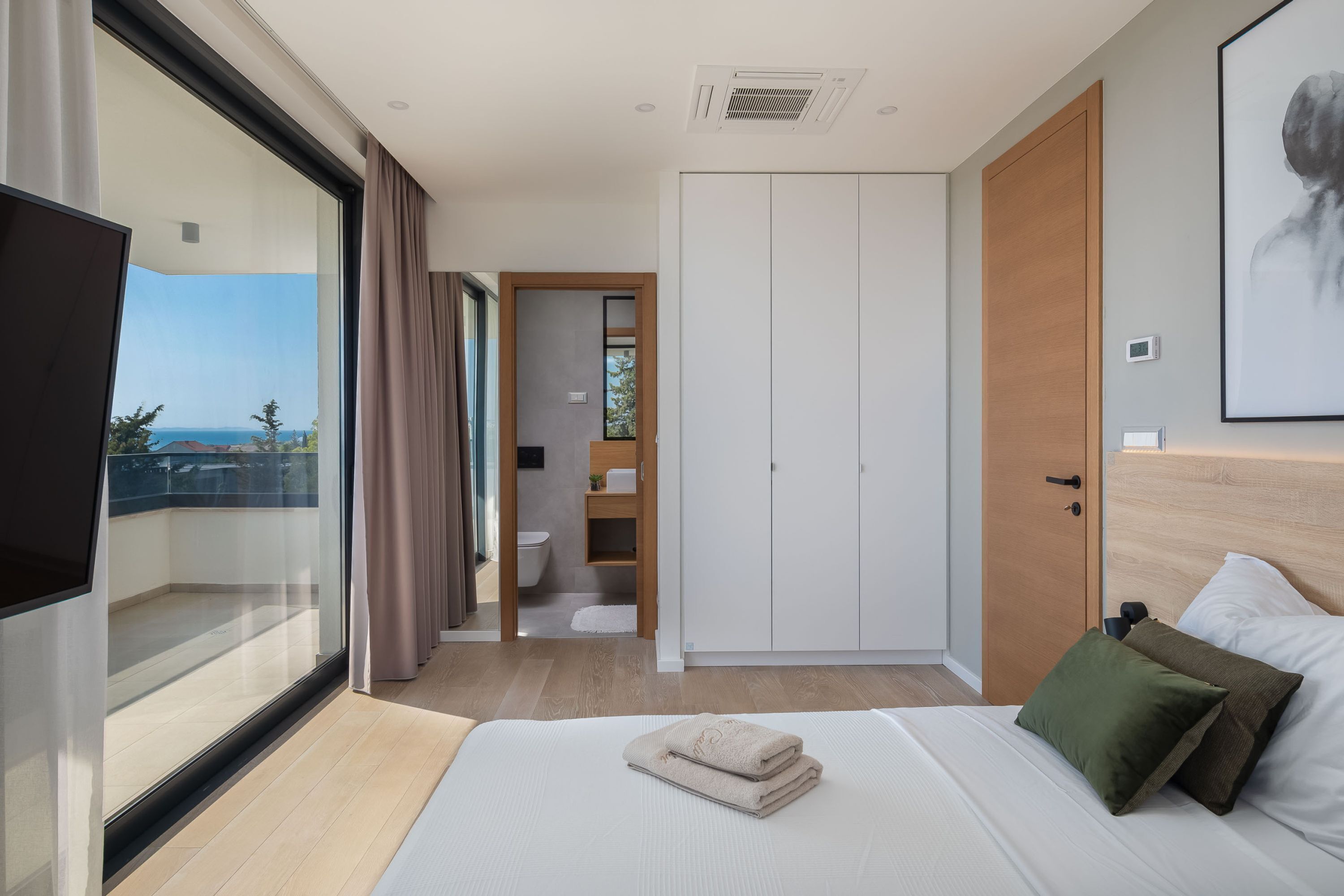 A modern hotel room with a large bed, a TV, a wardrobe, and a bathroom visible through an open door.