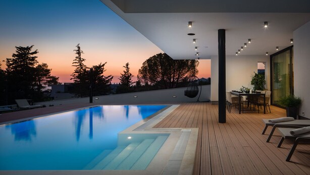 Un área de piscina moderna al aire libre con un deck de madera, una mesa de comedor con sillas y vistas a árboles contra un cielo al atardecer.