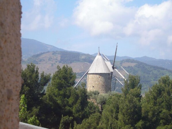 Eine steinerne Windmühle mit Metallverkleidung, umgeben von Bäumen und Hügeln im Hintergrund.