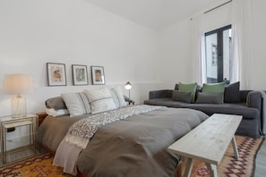 Un dormitorio moderno con cama, sofá, mesita de noche, lámpara y cuadros en la pared.