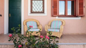 A building facade with a green door, a window with a metal grid, and two wicker chairs with striped cushions. - Casa Marzia by Wonderful Italy (Olbia)