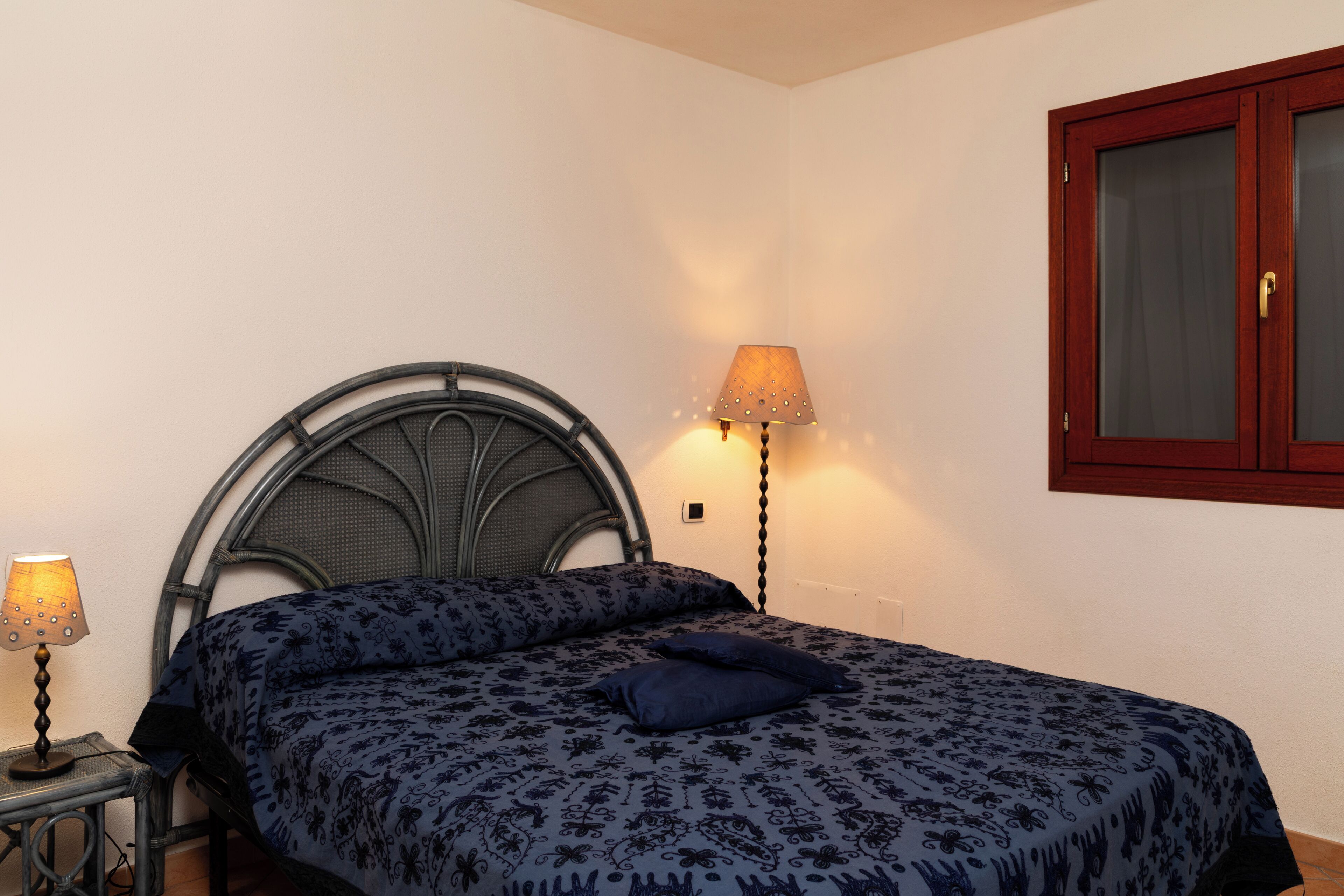 A bedroom with a bed, a nightstand, a lamp, and a window with a wooden frame.