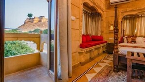 Een kamer met balkon met uitzicht op een fort, een bed met rode beddengoed en een traditionele houten stoel.