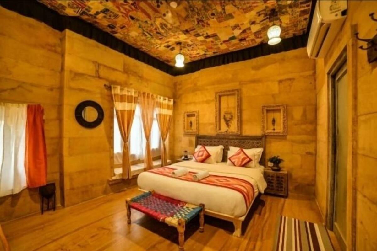 A room with a wooden floor, a bed with a colorful blanket, a bench, a wall-mounted clock, and a ceiling with a patterned design.