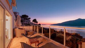 A balcony with lounge chairs and a table, overlooking a body of water and a mountainous landscape during sunset.