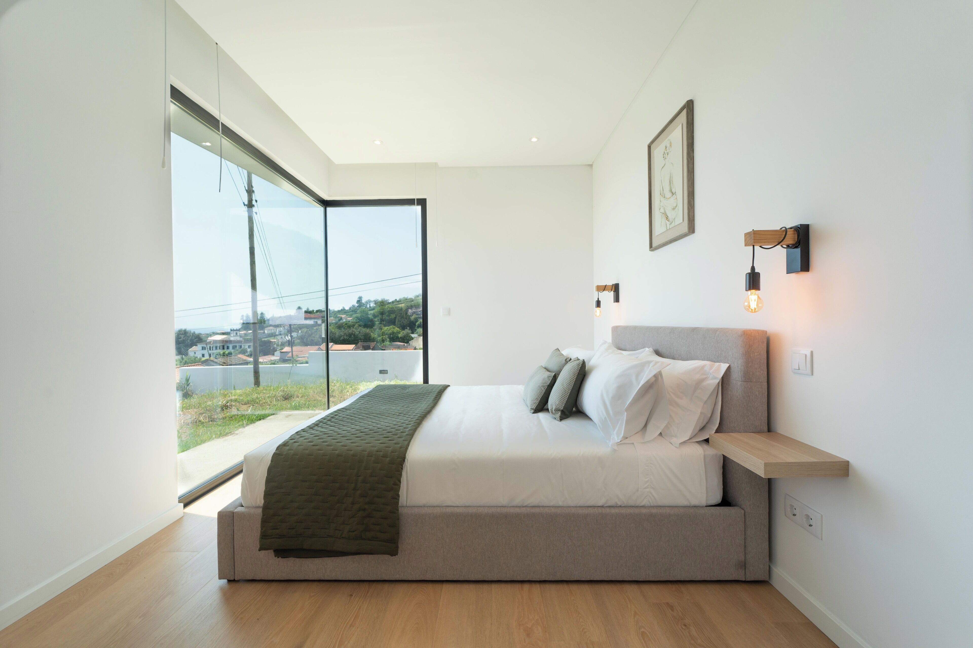 A modern bedroom with a large window, a bed with a wooden headboard, and a small bedside table.