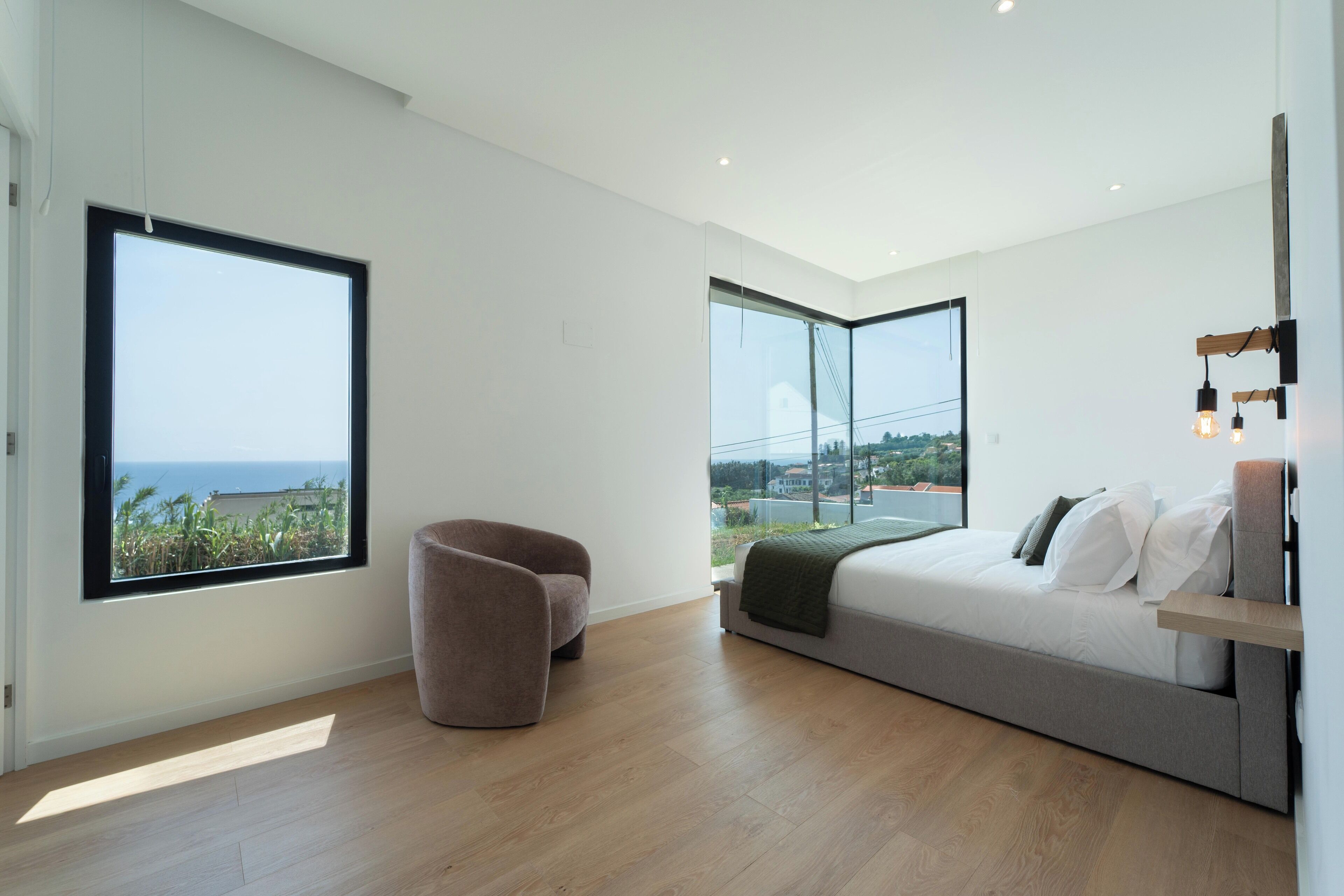 Ein modernes Schlafzimmer mit einem großen Bett, einem Sessel und zwei großen Fenstern mit Blick auf das Meer.