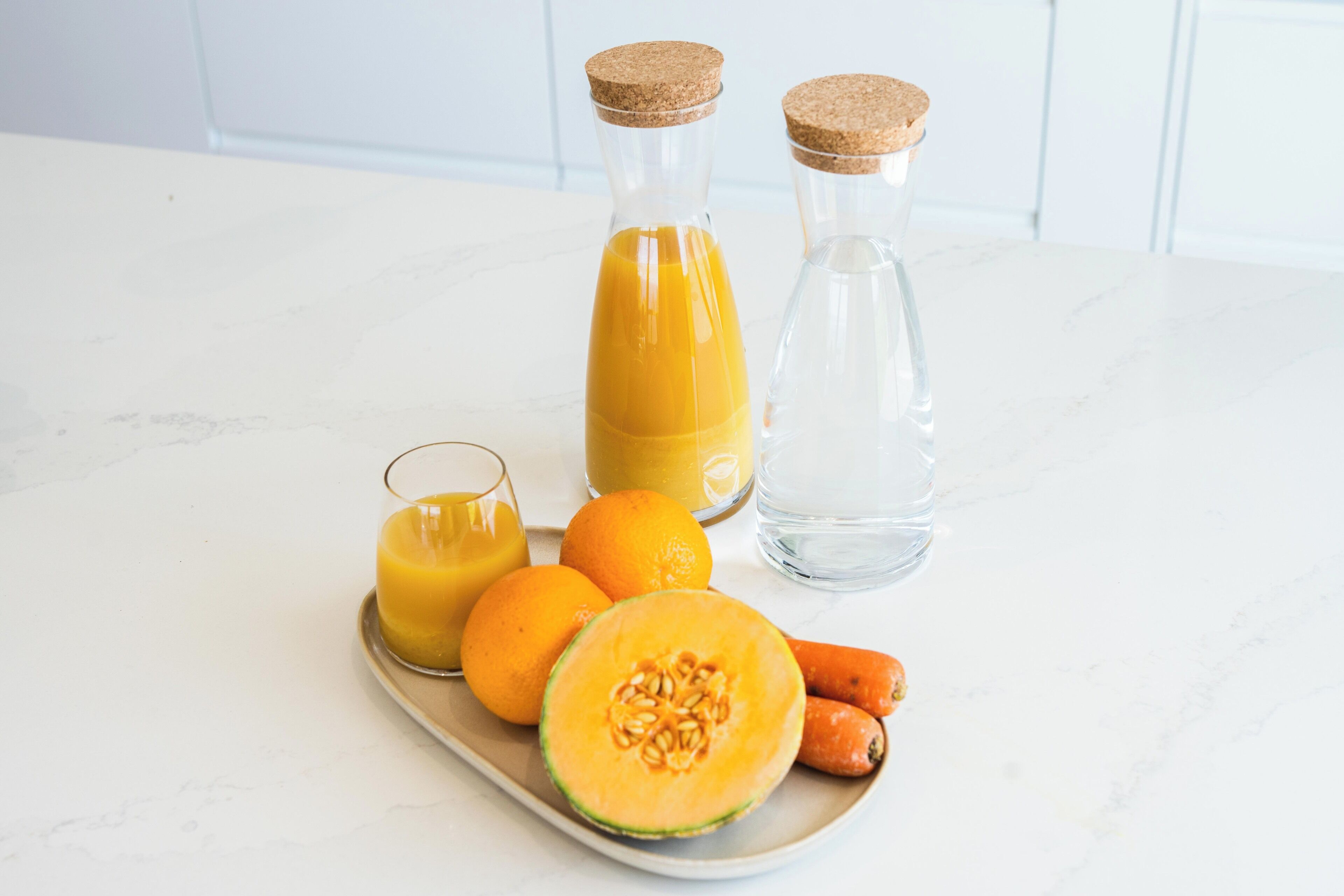 Eine Küchenarbeitsplatte mit einem Glas Orangensaft, zwei Orangen, einer halbierten Cantaloupe-Melone und zwei Wasserflaschen.