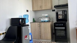 A compact kitchen with a refrigerator, microwave, coffee maker, and oven. There is a wooden cabinet above the sink and a chair with a table nearby.