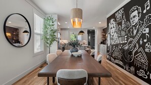 A dining area with a long wooden table, chairs, a round mirror, and a mural depicting Nashville music scenes. - New Stunning 4BR Nashville Home | Rooftop • Shuffleboard • Mural • Designer Interiors (Nashville)