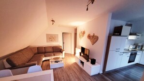 A modern living room with a sectional sofa, a flat-screen TV, and a kitchen area with white cabinets and a countertop. - Cozy vacation apartment with terrace and sauna (Röbel)