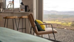 A chair with a cushion, a window with a view of the countryside, and a kitchen area with a coffee machine. - Migdale Heights - Luxury Cabins in The Highlands (Ardgay)