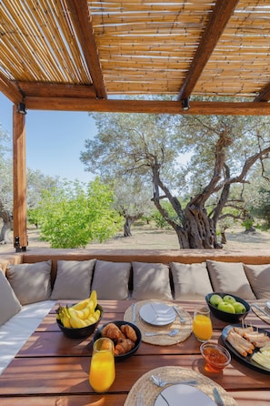 Un área de comedor al aire libre con una mesa dispuesta para el desayuno, que incluye frutas, croissants y jugo, bajo una pérgola de madera con vistas a árboles.