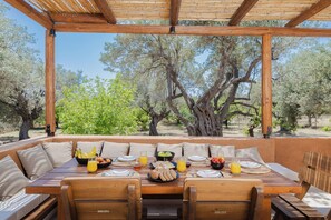 Un área de comedor al aire libre con una mesa dispuesta para el desayuno, rodeada de muebles de madera y vistas a olivos.