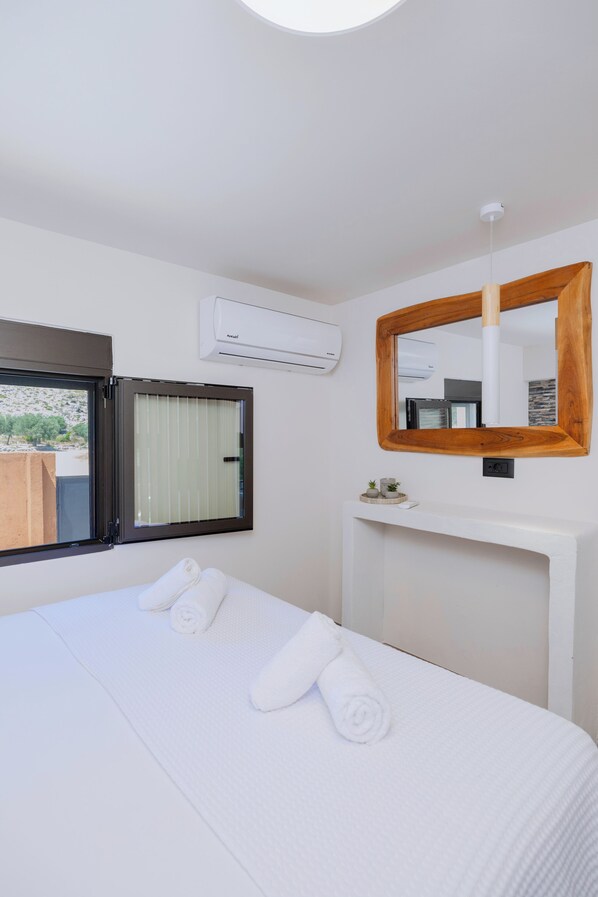 Habitación de hotel con cama, ventana, equipo de aire acondicionado, espejo y una pequeña estantería con objetos decorativos.