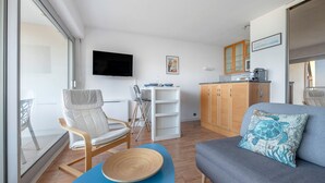 A modern living room with a grey sofa, a white chair, a blue table, and a kitchen area with wooden cabinets and a flat-screen TV. - Seafront, apartment for 4 people (Pornic)