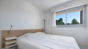 A modern bedroom with a wooden headboard, a white bed, and a window with a view of trees. - Seafront, apartment for 4 people (Pornic)