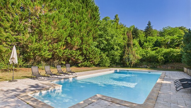 A rectangular swimming pool with a tiled deck, surrounded by lush greenery and trees.