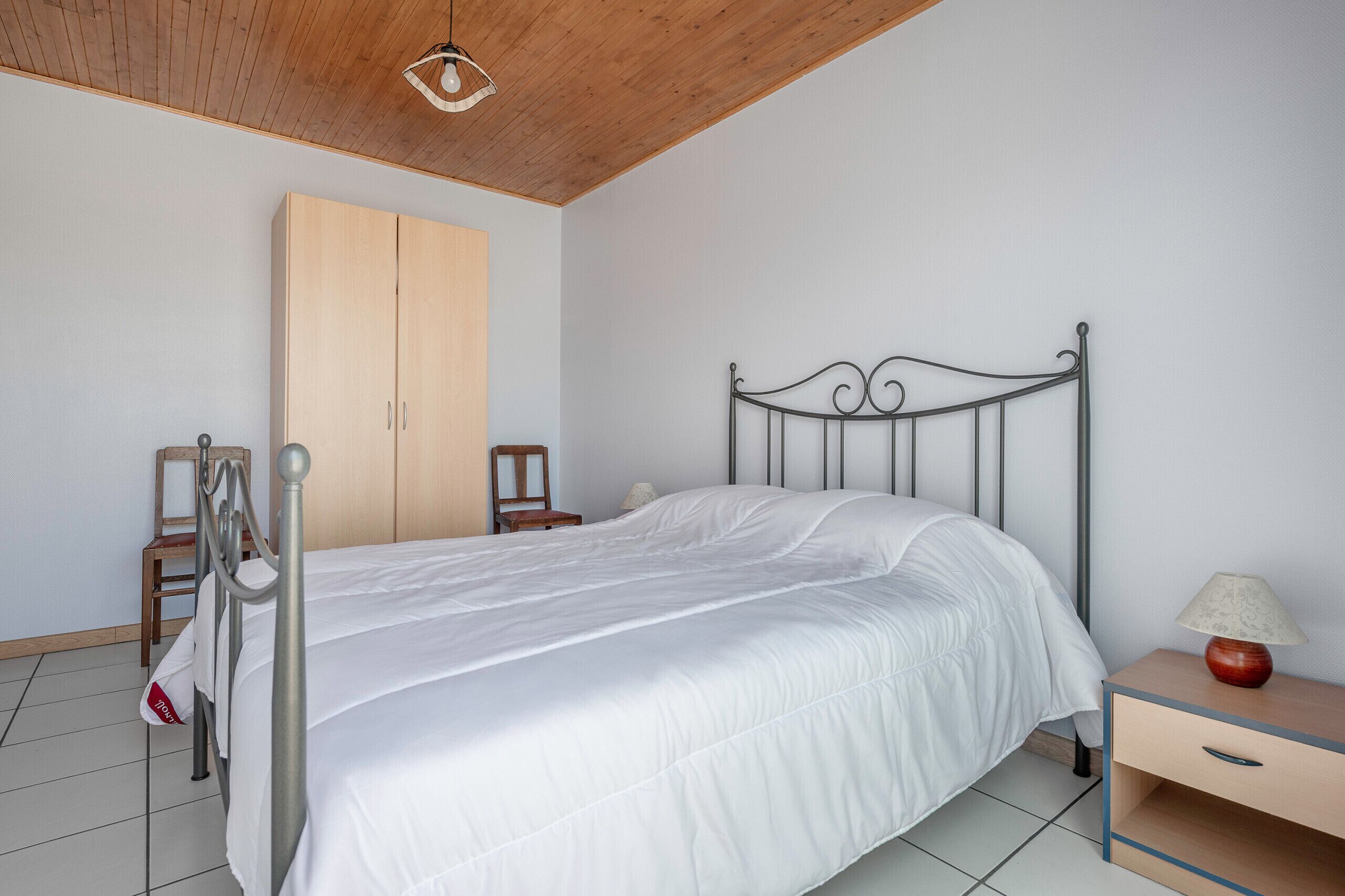 A bedroom with a metal bed frame, a wooden wardrobe, a bedside table with a lamp, and a chair.