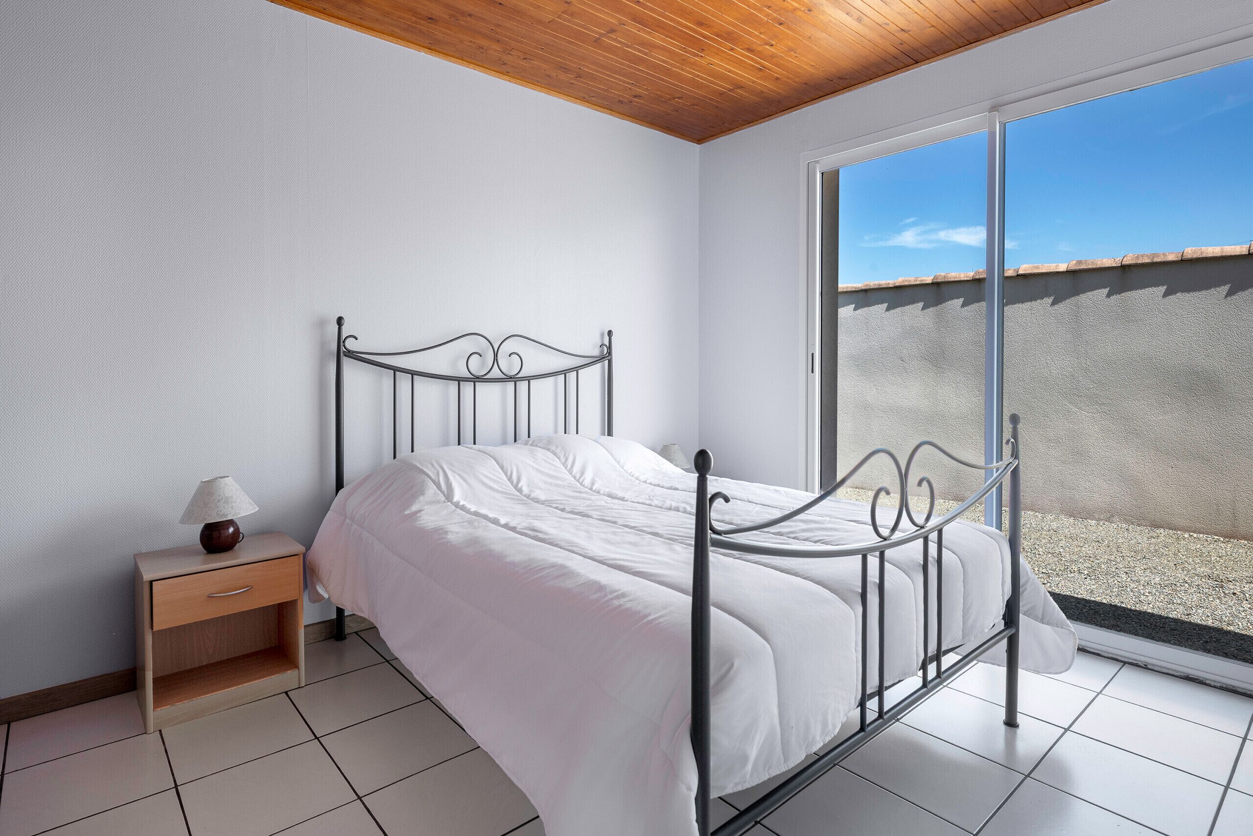 A bedroom with a metal bed frame, white bedding, a bedside table with a lamp, and a view of a balcony through a sliding door.