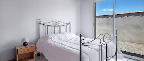 A bedroom with a metal bed frame, white bedding, a bedside table with a lamp, and a view of a balcony through a sliding door.