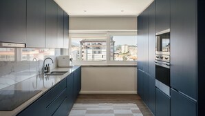 A modern kitchen with dark cabinetry, a marble countertop, a built-in oven, and a window with a city view.