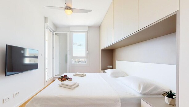 A modern hotel room with a large bed, a flat-screen TV, a ceiling fan, and a window with a city view. - Mika's 2-Bedroom Seaview Flat & Parking (Split)