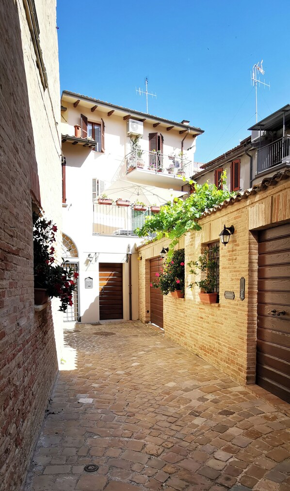 A narrow cobblestone alley with buildings on either side, featuring balconies with potted plants and a clear blue sky above.
