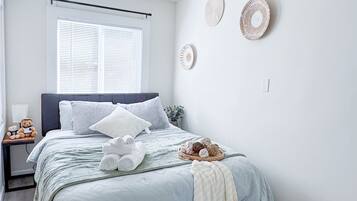 A neatly arranged bedroom with a bed, pillows, a bedside table with a teddy bear, and wall decorations.