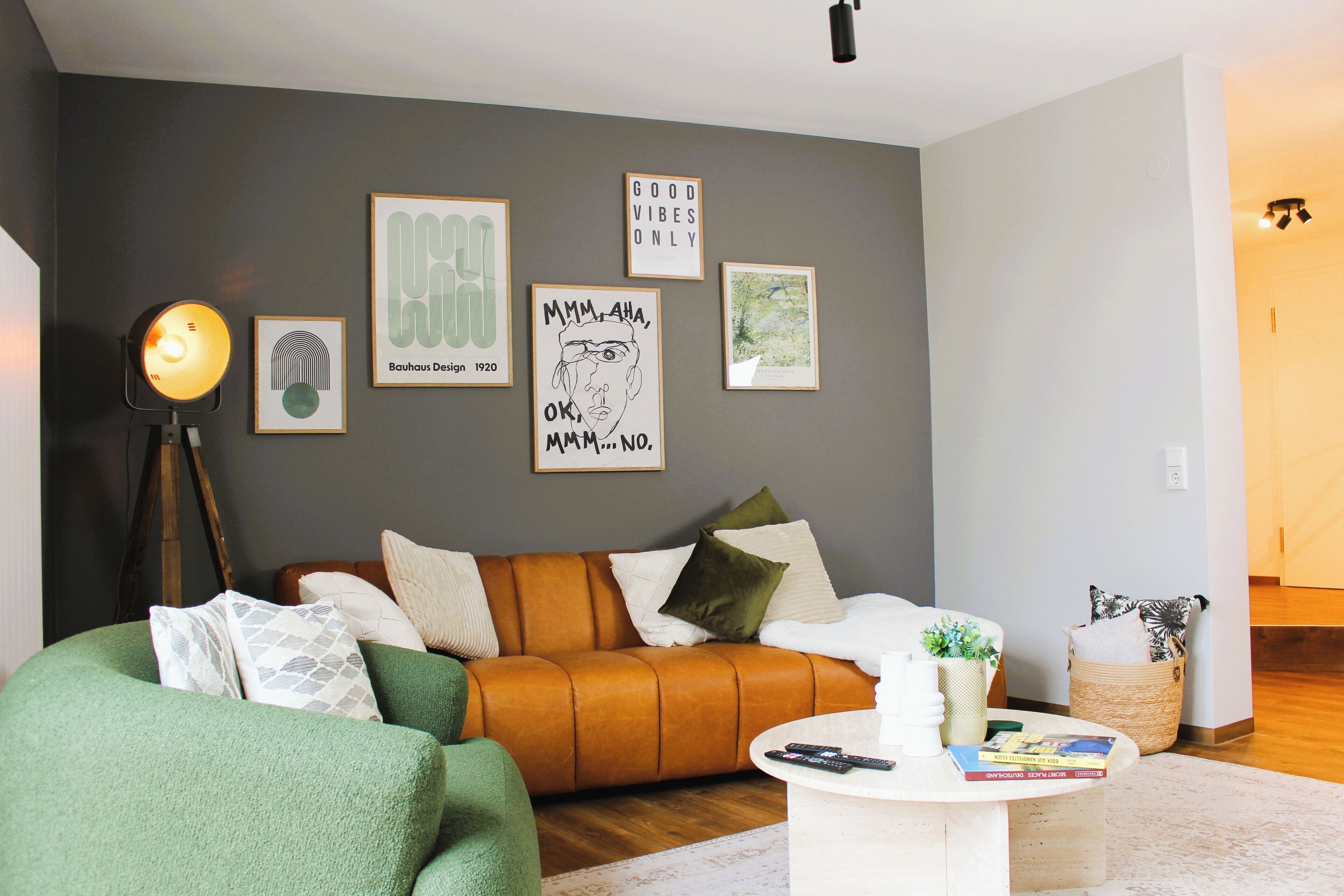 A living room with a green sofa, a brown leather couch, and a white coffee table. There are framed pictures on the wall and a floor lamp.