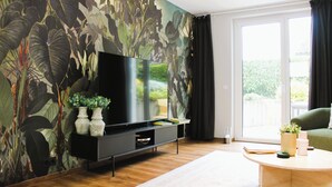 A living room with a large flat-screen TV on a black stand, a wooden floor, a green sofa, and a large window with curtains.