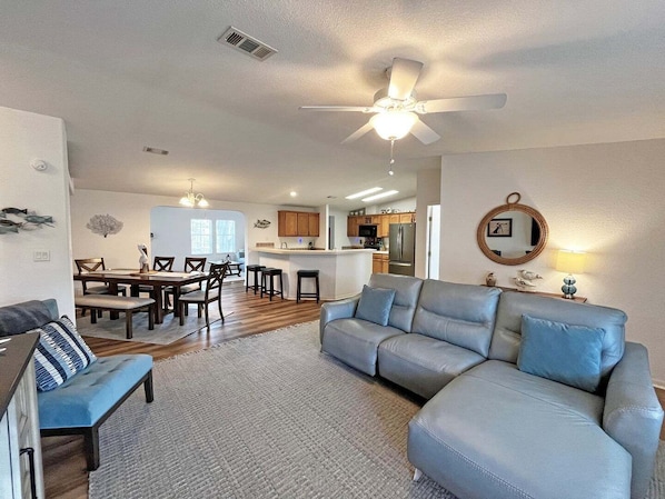 A living room with a gray sectional sofa, a dining area with wooden chairs, a kitchen with wooden cabinets, and a ceiling fan.