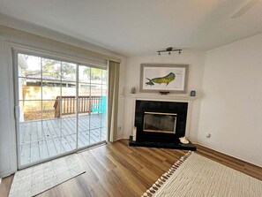 A room with a fireplace, a large window, and a wooden deck outside.