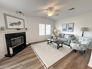 A living room with a fireplace, a ceiling fan, a sofa, a patterned armchair, and a coffee table.