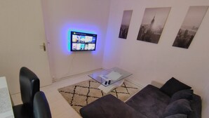 A modern living room with a flat-screen TV mounted on the wall, a glass coffee table, a grey sofa, and a black leather chair.