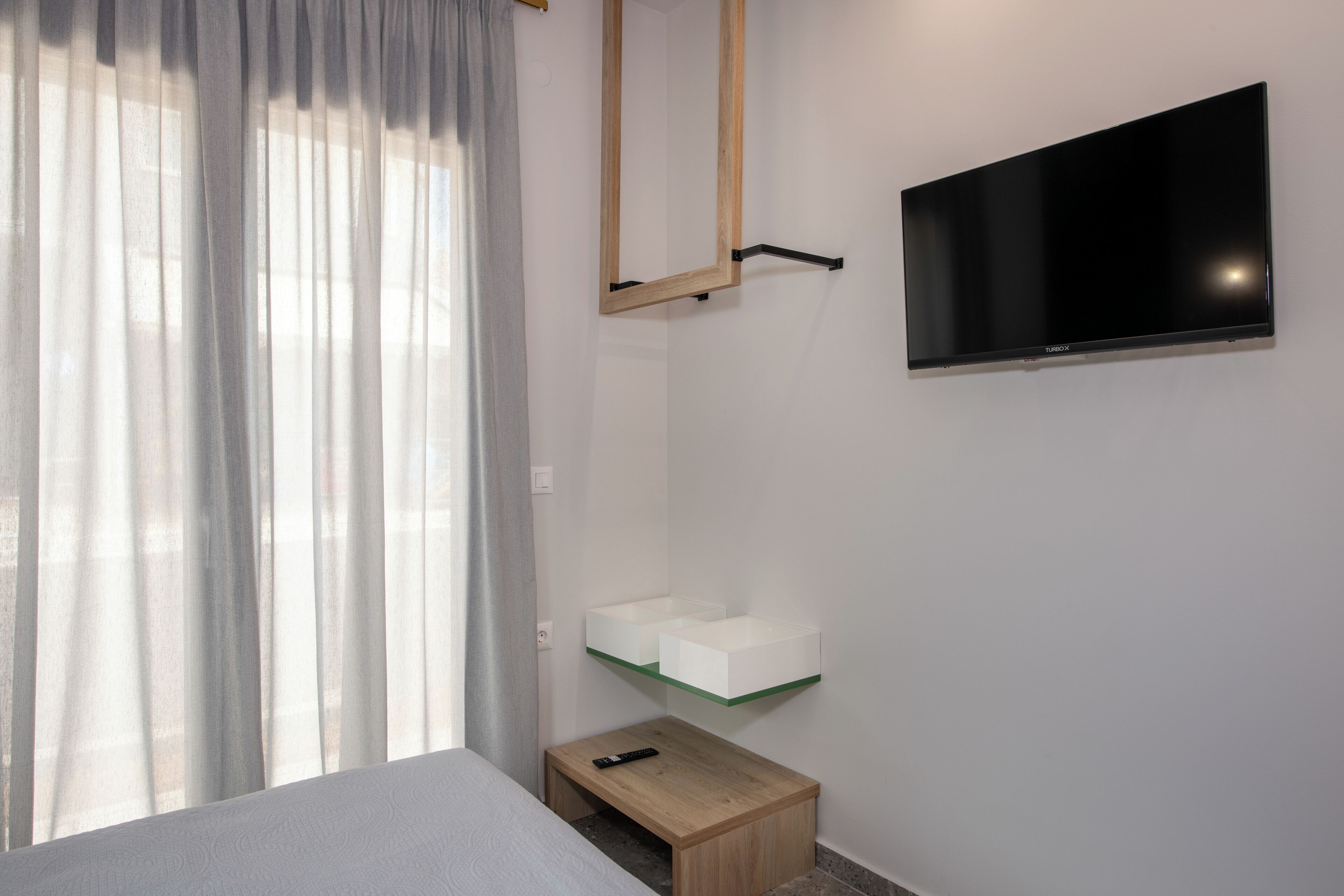 A modern hotel room with a bed, a mounted TV, a small wooden nightstand, and a glass shelf with two white bedside tables.
