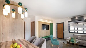 Ein modernes Wohnzimmer mit einem Holzespeisetisch, einer Obstschale, einem petrolfarbenen Sessel, einem goldfarbenen Couchtisch und einem Fernseher, der an einer grünen Akzentwand angebracht ist.