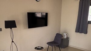 Hotellihuone, jossa on sänky, seinälle asennettu televisio, tuoli, pieni pöytä, jossa on vedenkeitin, ja ikkuna, jossa on verhot.