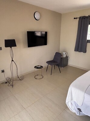 Una habitación de hotel con una cama, televisor montado en la pared, una silla, una mesita con un hervidor y una ventana con cortinas.